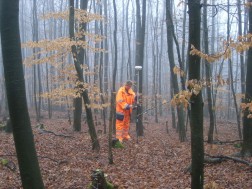 Eine Frau in orangefarbener Arbeitskleidung steht inmitten eines nebligen Waldes und hält eine lange Stange, an dem ein GPS-Meßgerät angebracht ist