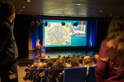 Foto vom hinteren Bereich eines vollen Veranstaltungssaals mit Blick auf die B&uuml;hne, wo gerade ein Vortrag gehalten wird