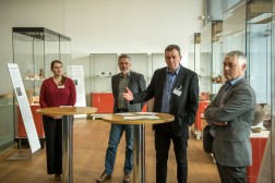 Vier Personen stehen an Stehtischen, eine Person gestikuliert, im Hintergrund die Vitrinen der Ausstellung.