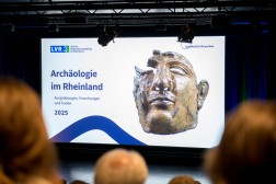 Blick auf eine per Beamer auf eine Leinwand projizierte Pr&auml;sentationsfolie auf einer B&uuml;hne, im Vordergrund sind unscharf Hinterk&ouml;pfe zu sehen