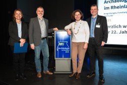 Vier Personen stehen auf einer B&uuml;hne um ein Rednerpult und l&auml;cheln in die Kamera