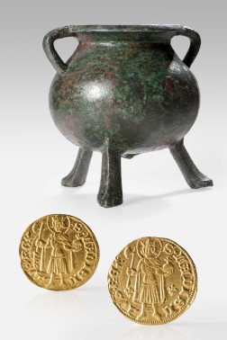 Ein grünliches Gefäß auf drei Füßen, im Vordergrund zwei goldene Münzen