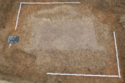Die ehemalige rechteckige Grabkammer mit Eckpfosten zeichnet sich deutlich in der kiesigen Anschüttung ab. Nur noch schlecht erkennbar ist der dunklere, sanduhrförmige Leichenschatten im Zentrum. Foto: Thomas Albert/LVR-Amt für Bodendenkmalpflege im Rheinland