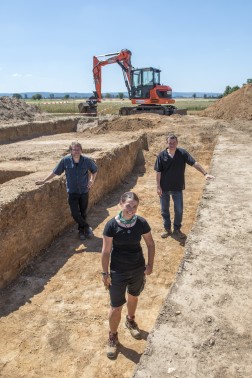 Eine Frau und zwei Männer stehen auf der Grabungsfläche und lächeln in die Kamera