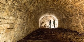Am Ende eines ausgeleuchteten Kellers mit gew&ouml;lbter Decke aus kleinen Felsquadern stehen zwei Personen vor einer Felswand und sprechen miteinander.