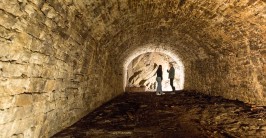 Am Ende eines ausgeleuchteten Kellers mit gew&ouml;lbter Decke aus kleinen Felsquadern stehen zwei Personen vor einer Felswand und sprechen miteinander.