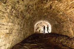 Am Ende eines ausgeleuchteten Kellers mit gew&ouml;lbter Decke aus kleinen Felsquadern stehen zwei Personen vor einer Felswand und sprechen miteinander.