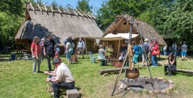 Mehrere Menschen stehen oder sitzen bei sonnigem Wetter auf gr&uuml;nem Rasen, im Hintergrund sind zwei H&auml;user mit gelblichem Lehmverputz und grau-braunem Strohdach.