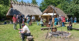 Mehrere Menschen stehen oder sitzen bei sonnigem Wetter auf gr&uuml;nem Rasen, im Hintergrund sind zwei H&auml;user mit gelblichem Lehmverputz und grau-braunem Strohdach.