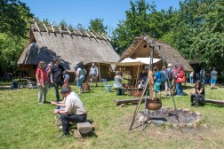 Mehrere Menschen stehen oder sitzen bei sonnigem Wetter auf gr&uuml;nem Rasen, im Hintergrund sind zwei H&auml;user mit gelblichem Lehmverputz und grau-braunem Strohdach.