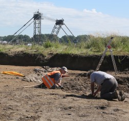 Zwei Personen knien an einer Grube, dahinter sind Teile eines großen Baggers zu sehen.
