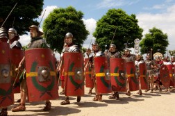Mehrere m&auml;nnliche Reenactor laufen bei sonnigem Wetter in r&ouml;mischer R&uuml;stung mit roten Schilden in einer Reihe hintereinander