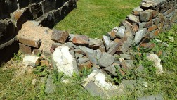 Eine kleine Mauer aus losen grauen Steinen, wo in der Mitte die Steine heruntergefallen sind und vor der Mauer auf dem grünen Gras liegen.