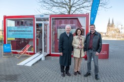 Drei Personen stehen vor einem roten Glascontainer und l&auml;cheln in die Kamera, im Hintergrund ist in der Ferne der K&ouml;lner Dom zu erkennen.