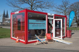 Foto des Glascontainers mit rotem Rahmen bei sonnigem Wetter, im Hintergrund ist in der Ferne der K&ouml;lner Dom zu erkennen.